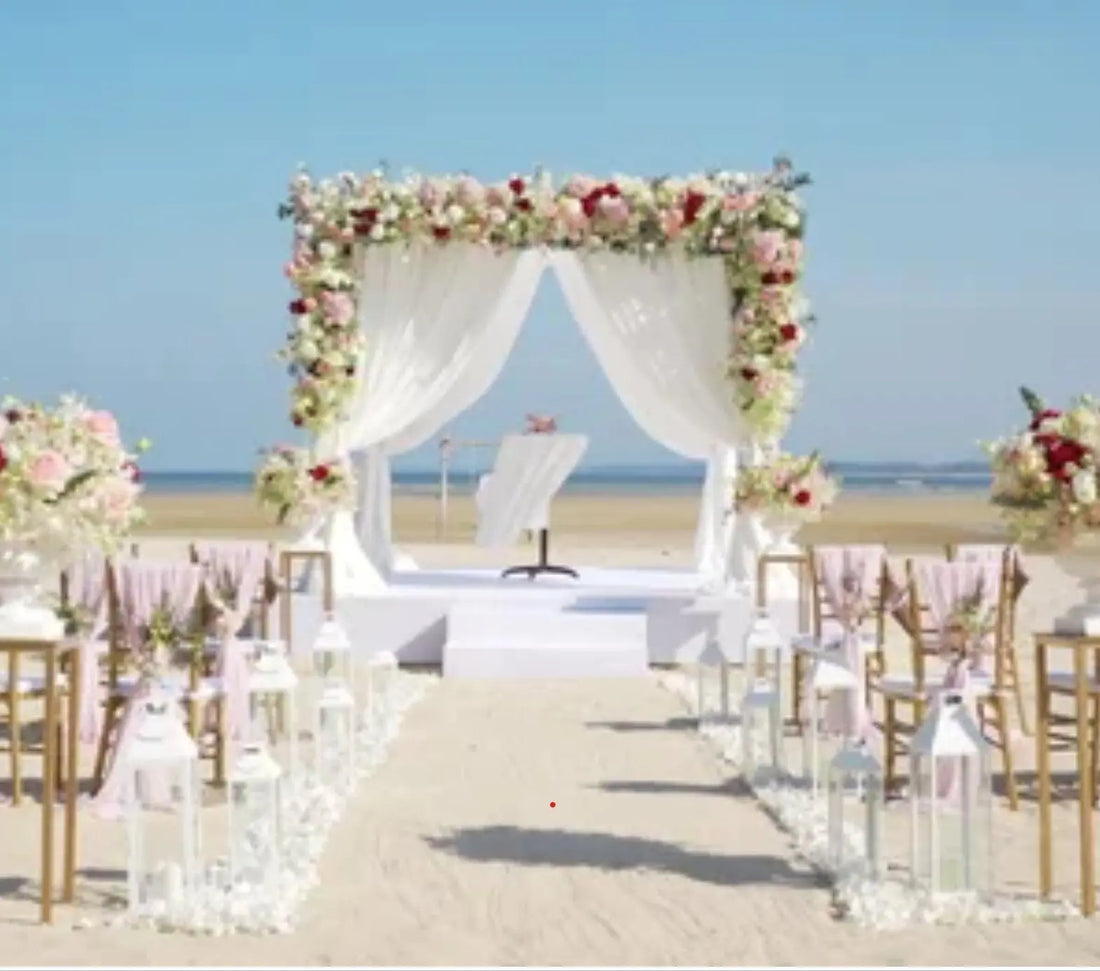 Beautiful beach wedding ceremony setup with a white floral arch, draped curtains, pastel flowers, and lanterns lining the sandy aisle under a clear blue sky