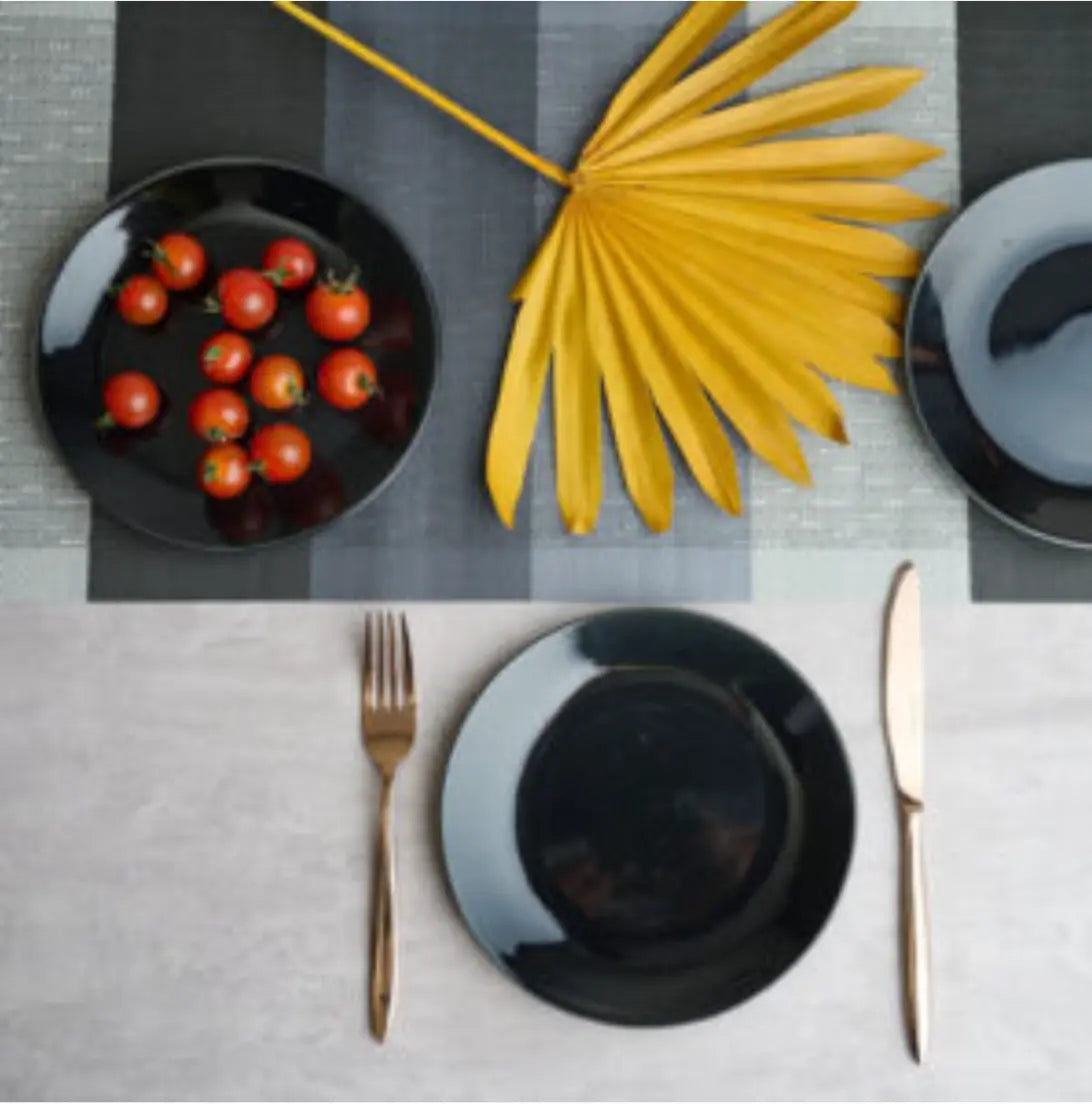 modern table setting with black ceramic dinner and salad plates, gold cutlery, and cherry tomatoes on a checkered placemat with a yellow dried palm leaf