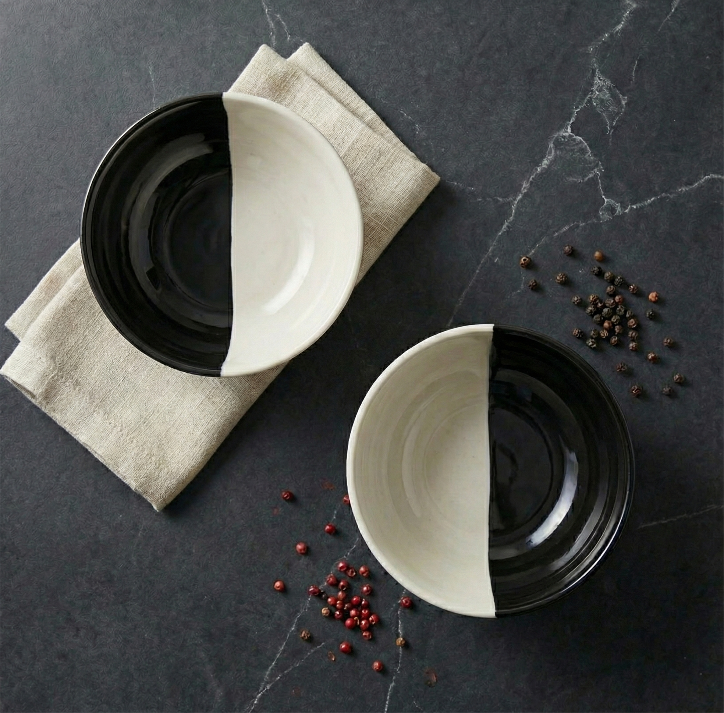 Two ceramic bowls, one black and one white, on a dark marble surface with scattered peppercorns.